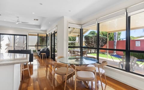 A modern, well-lit living space with a wooden floor and large windows offering a view of the garden outside. It features a glass dining table with wooden chairs, a cozy seating area with sofas, and a clean, bright interior design that creates an inviting 