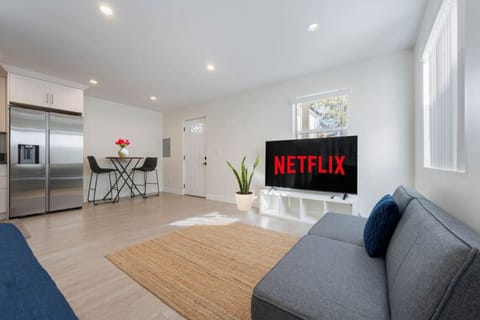 A cozy living area with a gray sofa bed, large TV ready for Netflix, a small dining nook, and a stainless steel fridge.