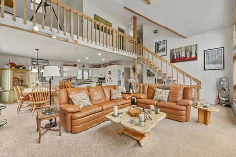 Open-concept living room with a leather sectional and coffee table, seamlessly flowing into the kitchen and dining area beneath a lofted staircase.