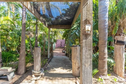A rustic wooden walkway surrounded by lush tropical plants and trees, with a canopy providing shade. The walkway features weathered wooden posts and a quaint lantern, creating a charming and natural garden pathway.