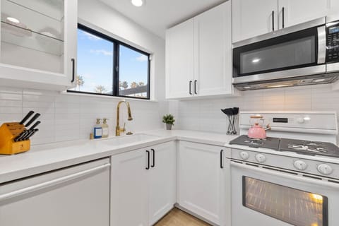 A modern and compact kitchen corner, featuring crisp white cabinetry and subway tile backsplash. The space is equipped with a stainless steel microwave, oven, and a stove, accented by a stylish gold faucet and matching handles.
