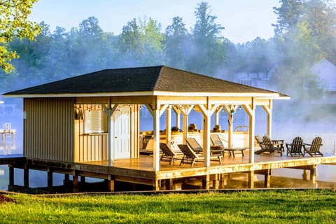 A picturesque covered boathouse and dock featuring lounge chairs, a pontoon boat slip, and Adirondack seating, framed by mist rising off the lake at sunrise.