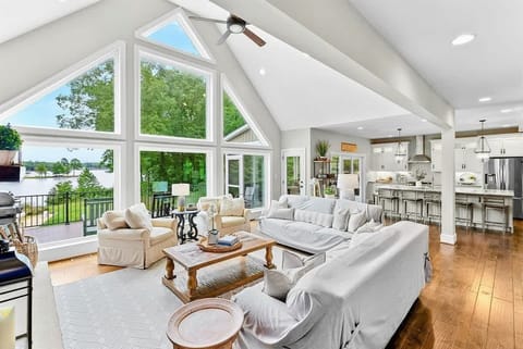 A bright and spacious lakefront living area with large floor-to-ceiling windows, cozy seating, a stone fireplace, and open access to the kitchen and outdoor deck.