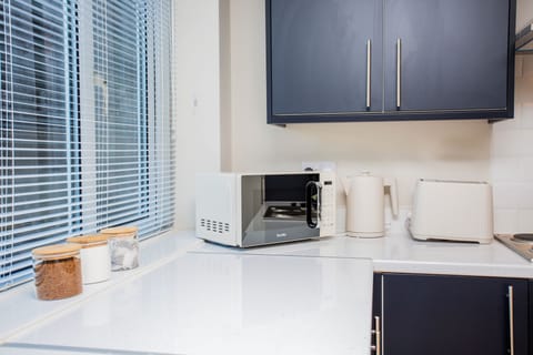 This image shows a section of the kitchen with a microwave, a kettle, and a toaster on a clean countertop. Two glass jars containing dry ingredients are visible next to the appliances. The cabinets are dark blue, and there’s a window with blinds allowing natural light to enter. The white countertop contrasts well with the blue cabinets.