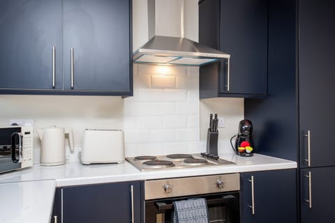 This image showcases a well-equipped kitchen with navy blue cabinets, a stainless steel stove, and modern appliances, including a microwave, toaster, and coffee machine. The kitchen is accented with a white tiled backsplash and a tea towel hanging from the oven.