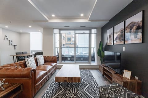 `Stylish open living room featuring a classic tufted leather Chesterfield sofa, dark accent wall with framed artwork, flat-screen TV, patterned area rug, and sliding glass doors to the balcony.`