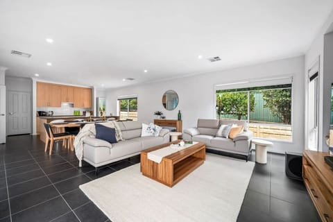 A modern, open-plan living space with a sleek design. It features light-colored sofas adorned with cushions, a wooden coffee table, and a light-colored rug on dark tiled flooring. The area includes a kitchen with wooden cabinets in the background and larg