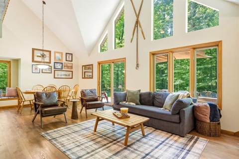 Bright, open-concept living and dining area with soaring ceilings, floor-to-ceiling windows, and peaceful forest views that bring the outdoors in.