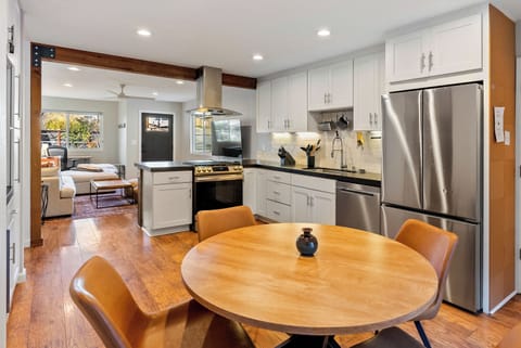 Step into this bright, fully stocked kitchen and dining space, featuring sleek white cabinetry and a warm wooden dining set. The open layout flows seamlessly into a cozy living area, creating the perfect environment for gathering and entertaining in style.