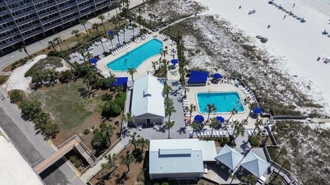 This aerial view captures the resort's stunning beachfront amenities, featuring two sparkling swimming pools, spacious sundecks with loungers, shaded seating areas, and direct access to the sugar-white sands of the Gulf. Surrounded by palm trees and