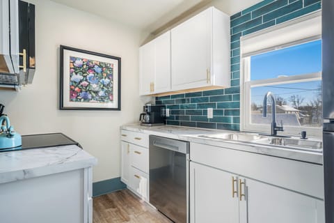 Cook in style in this modern kitchen with sleek white cabinetry and gold hardware. The space is brightened by a large window and features a marble countertop with a deep sink. Stainless steel appliances include a dishwasher and coffee maker.