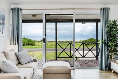 A cozy living room with a light-colored sofa and ottoman, complemented by light gray curtains. Through a large sliding door, there is a view of a wooden deck with a railing and an outdoor scene of grassy land under a cloudy sky, suggesting a serene and co