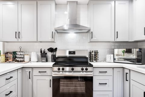 Cook with ease in this fully equipped and modern kitchen! The centerpiece is a sleek stainless steel oven with an overhead range hood, surrounded by stylish gray cabinets and a pristine white tile backsplash.