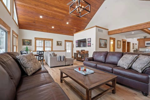Scott Creek Lodge living room with sectional & 60" TV + small games table. To the left - back deck access; behind you - fireplace & screened porch access; to the right - kitchen; straight ahead, near the main entrance door - basement stairs.