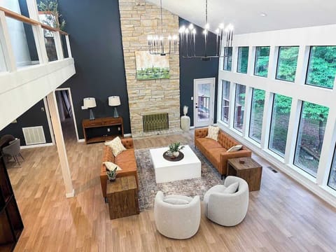 Relax in this grand, light-filled living room featuring vaulted ceilings, floor-to-ceiling windows, a stone accent fireplace wall, cognac leather sofas, rounded accent chairs, and an upper-level loft view.