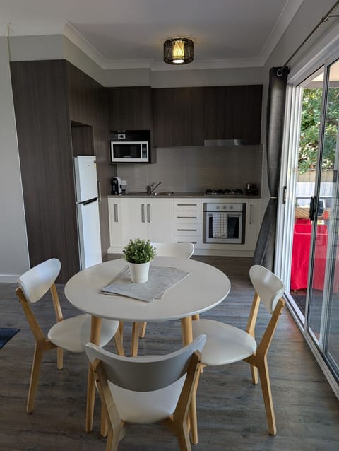A modern kitchen and dining area with dark wood cabinets, a white refrigerator, and a stainless steel oven. There is a round dining table with four white chairs and a potted plant on it, situated near a sliding door that offers a view of greenery outside.
