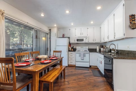 Step into a bright, inviting kitchen and dining area where modern comforts meet rustic charm, featuring white cabinetry, granite countertops, and a spacious wooden table perfect for gathering with family and friends. Floor-to-ceiling windows and sliding doors bathe the space in natural light, seamlessly connecting you to the serene outdoor surroundings for a truly relaxing stay.