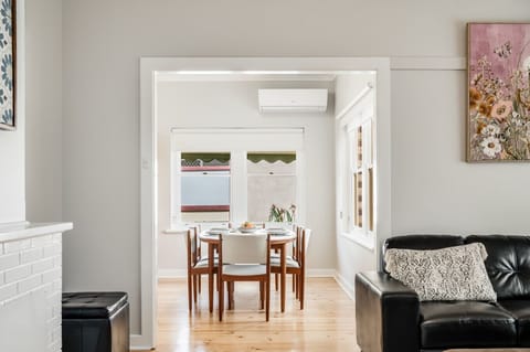 A modern, stylish home interior with a light-colored wall and wooden floor. There is a dining area with a round table and chairs visible through an open doorway, and a living room area with a black leather sofa adorned with a patterned cushion. The decor 