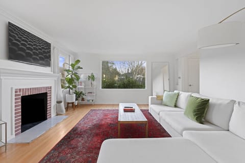 A flat-screen TV is mounted above the fireplace, and a tall potted plant adds a touch of greenery. The room is well-lit with natural light from a large picture window and smaller side windows, offering a view of the outdoors.