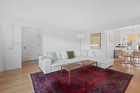 A large white sectional sofa with green accent pillows provides plenty of seating, complemented by a wooden coffee table over a vibrant red Persian-style rug.