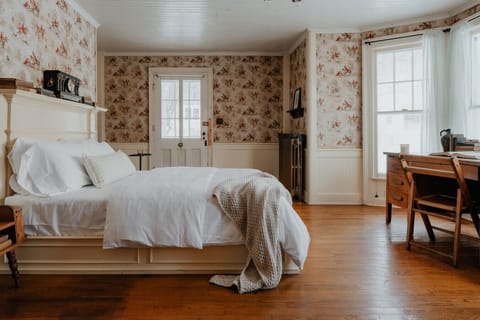 Spacious and Elegant Sleeping Area – A bright and inviting bedroom featuring a grand wooden bed, crisp white linens, and vintage-inspired décor. The room is bathed in natural light from large windows, creating a serene and comfortable atmosphere.