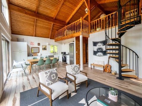 Bright open-concept living room with vaulted wood ceilings, spiral staircase to loft, two accent armchairs, and a view toward the dining area for 8 and kitchen.