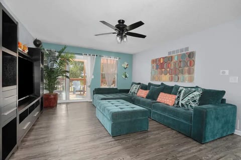 This stylish Airbnb features a spacious living area with a plush teal sectional sofa, vibrant decor, and a ceiling fan. Large windows invite natural light, creating a warm and inviting atmosphere for relaxation.