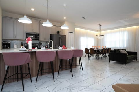 Sleek kitchen flows to a sunlit dining area and cozy lounge-quartz island with pink stools, a sparkling chandelier, and airy drapery create a modern-glam setting for gathering.