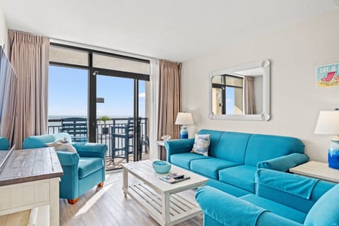 Bright, oceanfront living room featuring a full sofa and queen sofa sleeper, a 65” Vizio Smart TV, and floor-to-ceiling windows. Natural light pours in, highlighting stunning ocean views and direct access to the private balcony.