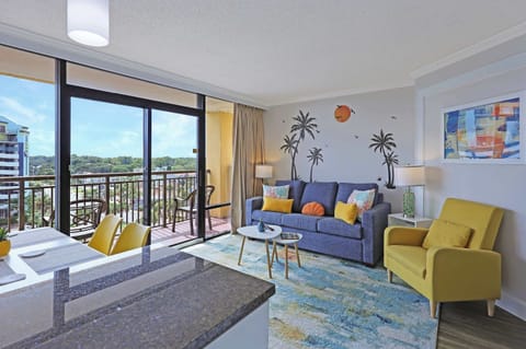 Relax in this sunlit living room with tropical wall art, cozy seating, and balcony access for ocean breezes and city views—your perfect spot to unwind after a day at the beach.