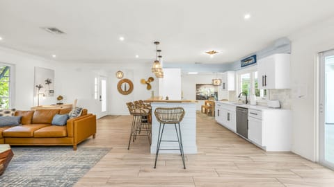 The bright and airy living room features a modern leather sectional, warm wood accents, and lush greenery views through large picture windows. Stylish touches like the surfboard décor and geometric coffee table create a relaxed coastal vibe.