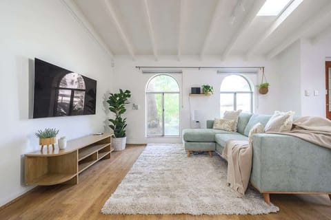 Spacious and serene living room with a huge, comfy sofa, large smart TV, and abundant natural light. Unwind in this bright, plant-filled haven—your perfect home away from home!