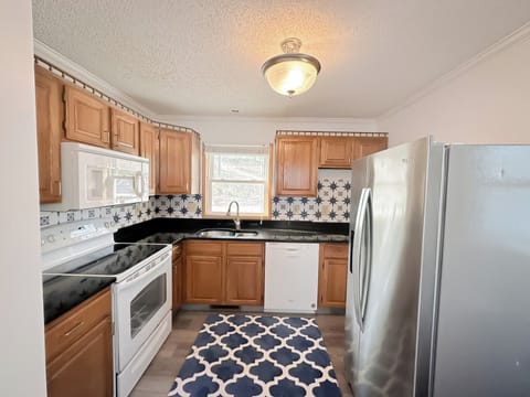 This well-equipped kitchen features warm wood cabinetry, sleek black countertops, and modern stainless steel appliances including a refrigerator, microwave, dishwasher, and electric range. The stylish blue and white patterned rug adds a touch of charm to the light wood-look flooring, creating a functional and inviting space for meal preparation.