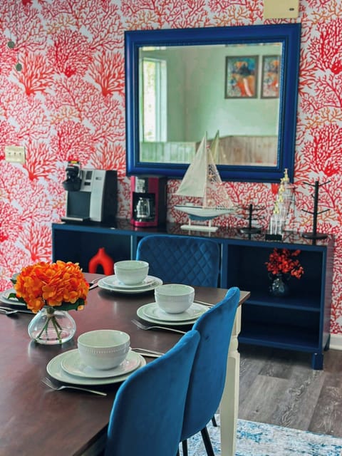 This vibrant dining area features a rich wooden table surrounded by stylish blue velvet chairs, creating an inviting space for meals. The eye-catching coral-themed wallpaper and a striking blue sideboard with nautical accents add a unique and cheerful coastal charm to the room.