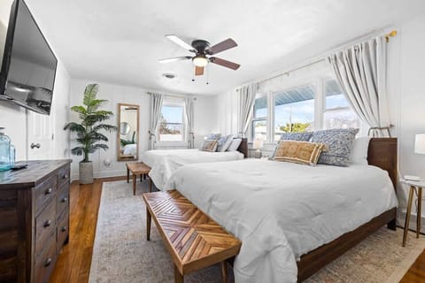 A spacious master bedroom features a large bed, bright natural light, and elegant color. The airy curtains and ceiling fan enhance the relaxing vibe.