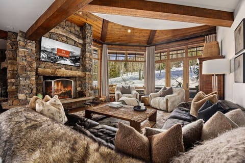 View of a luxury living room with ski slopes visible through a wall of windows. The couch is large and overstuffed with many pillows. there are two faux fur covered couches, a large fireplace and a TV