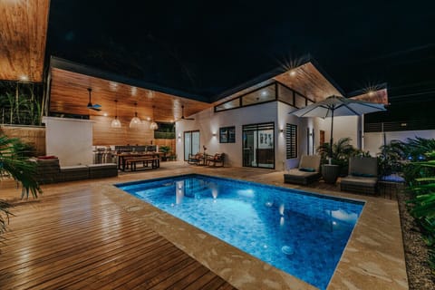 A dreamy nighttime view of this modern tropical villa—crystal blue pool, glowing lights, a cozy rancho with plush seating, BBQ grill, outdoor kitchenette with stove, sink, and beverage fridge, perfect for unforgettable open-air dining.