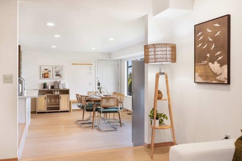 Bright open-concept dining and lounge area with a Scandinavian-inspired wooden table, green-cushioned chairs, and a woven floor lamp beneath avian wall art—where natural light meets effortless elegance.