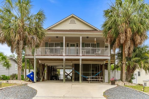 Raised beach home framed by palm trees with covered parking, hammock lounge, wraparound porch seating, and spacious driveway in a quiet coastal neighborhood.