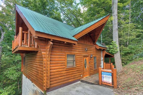 Charming log cabin with green metal roof nestled among towering trees, featuring a welcoming entrance and rustic mountain architecture.