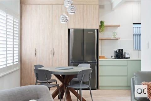 A modern kitchen and dining area with a sleek design. It features light wood cabinetry, a stainless steel refrigerator, a round dining table with perforated chairs, stylish geometric pendant lights, and light green lower cabinets. The space also includes 