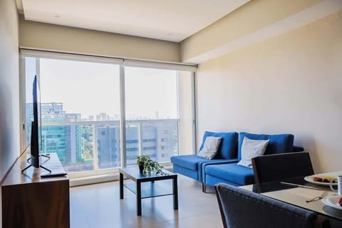 Sunlit living area w/ sofa seating, TV & floor-to-ceiling windows framing sweeping city views