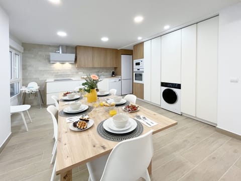 Open kitchen with white and wood cabinets, a dining area with a light wood table and six chairs, with a washing machine on the right.