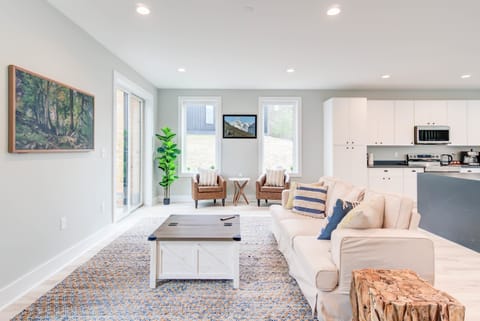 A cozy living room with two side chairs and a small table, complemented by a kitchen bathed in natural light from the glass window.