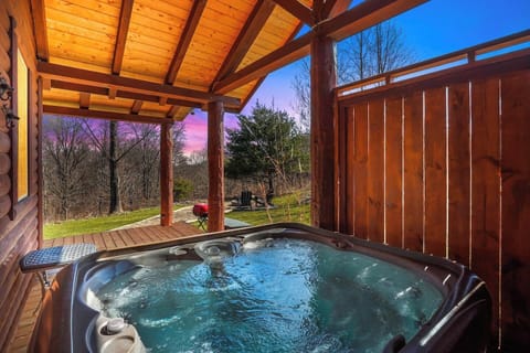 Unwind in the bubbling hot tub on the covered deck at My Little Heaven, surrounded by warm wood accents and tranquil views—perfect for relaxing after a day of exploring or savoring a quiet evening as the sun sets over the trees.