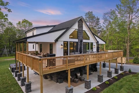 The modern farmhouse exterior glows at dusk, with a wraparound deck ready for gathering.
