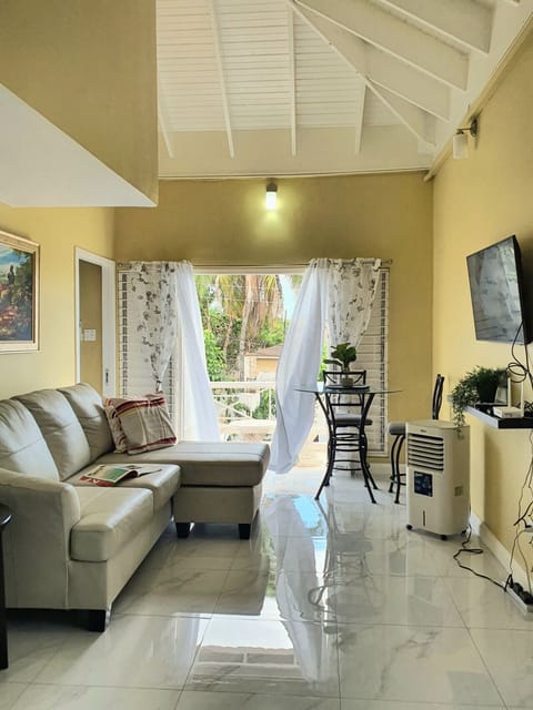 Bright living area w/ sofa & TV, opening to a breezy balcony through sheer curtains.