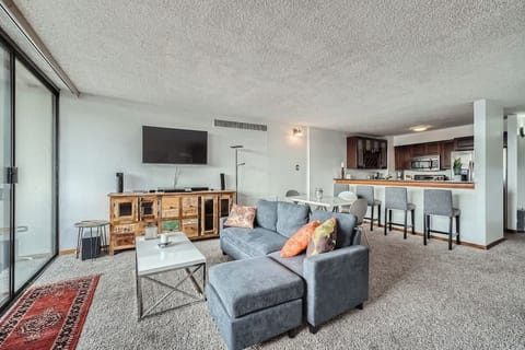 Bright and modern open-plan kitchen flowing seamlessly into a cozy living area with natural light. Features include bar tops and full-size appliances, perfect for cooking or gathering.