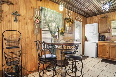 A rustic dining area and kitchen space featuring wood-paneled walls, a corrugated metal ceiling, a charming wrought iron dining set, and a stacked washer and dryer unit, all adorned with country-themed decor.