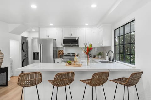This sunlit kitchen boasts stainless steel appliances, rattan bar seating, and sleek white cabinetry. A picture-perfect spot for morning coffee in your Crown Point Airbnb—just a short walk to the bay and all things San Diego.
